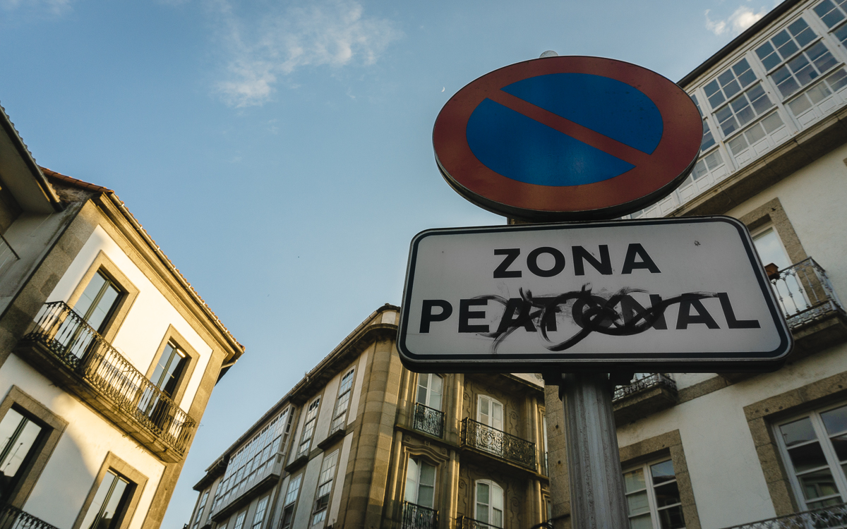Señal de zona peatonal tachada con graffiti con edificios de la calle de Porta do Camiño de fondo.